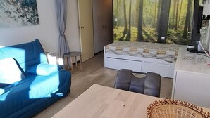 A modern living room with a sofa, a coffee table, a bed with a forest mural, and a kitchen area with a stove. - Ski-in/ski-out & foot of the slopes, 6th floor, balcony, tv, 24m², La Plagne (La Plagne)