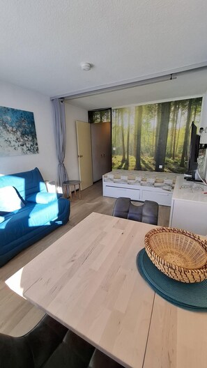 Ein modernes Wohnzimmer mit Sofa, Couchtisch, einem Bett mit Waldmotiv und einer Küchenzeile mit Herd.