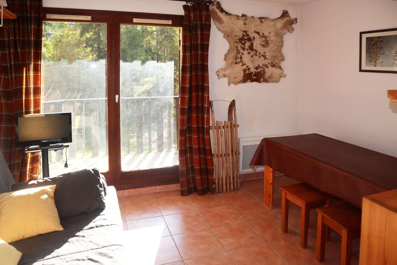 A room with a sofa, a television on a stand, a dining table with benches, a fireplace, and a large window with curtains.