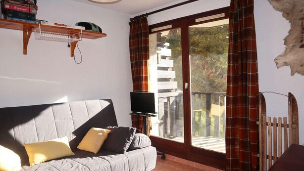 Un salon avec un canapé gris, un téléviseur à écran plat, une étagère en bois remplie de livres et un balcon agrémenté de rideaux à carreaux rouges.