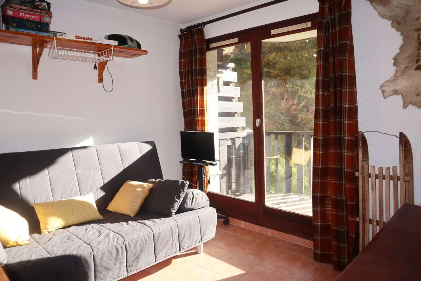 A living room with a grey sofa, a flat-screen TV, a wooden shelf with books, and a balcony with red plaid curtains.