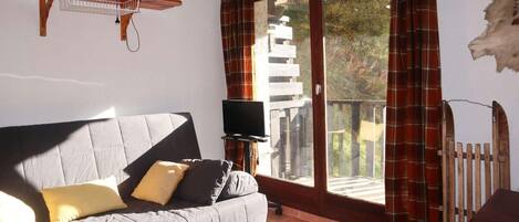 A living room with a grey sofa, a flat-screen TV, a wooden shelf with books, and a balcony with red plaid curtains.