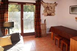 A room with a sofa, a television on a stand, a dining table with benches, a fireplace, and a large window with curtains.