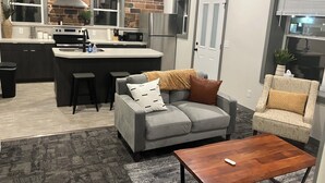 A modern kitchen with a central island, bar stools, a grey sofa, a wooden coffee table, and a patterned armchair.