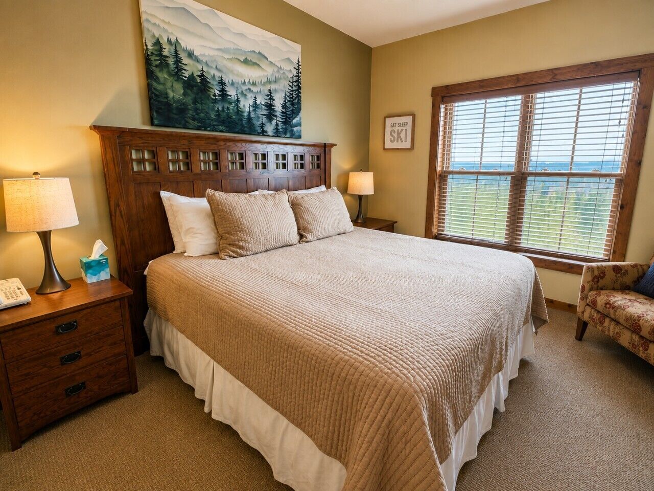 A bedroom with a large bed, wooden headboard, two bedside lamps, a nightstand with a phone, a chair, and a window with blinds.