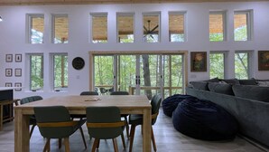 Et moderne interiør med et træbord, stole, en grå sofa og et stort vindue med udsigt til træer.