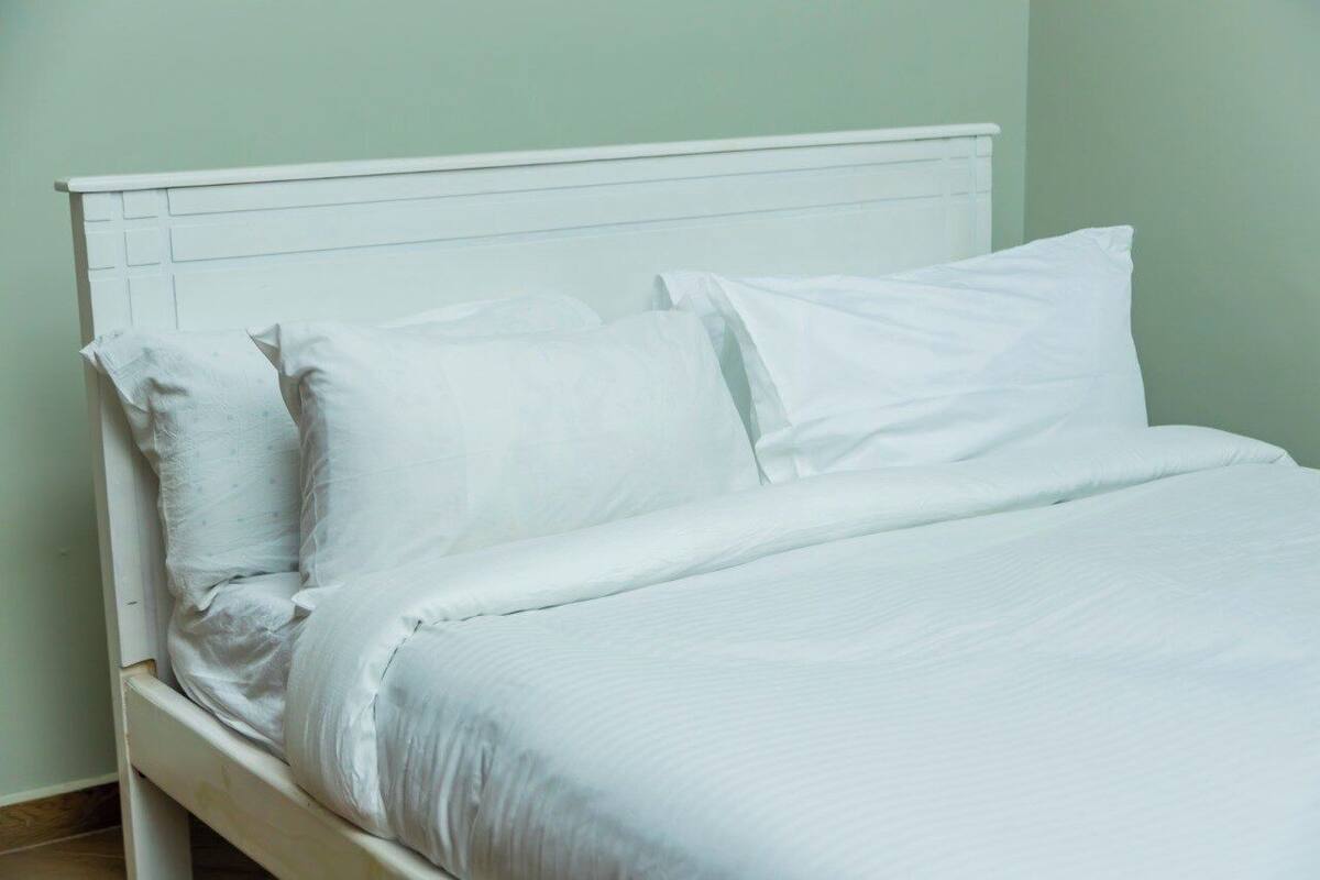 A neatly made bed with white linens and pillows in a room with light green walls and a white headboard.
