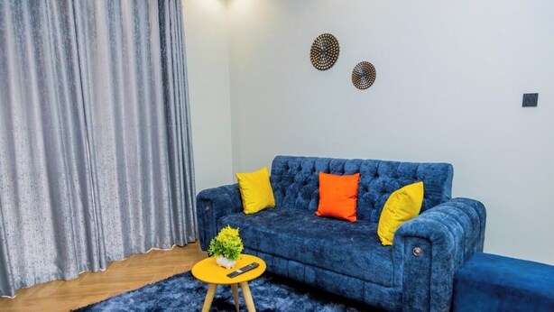 A living room with a blue sofa, yellow and orange cushions, a small round yellow table with a plant and remote control, a blue furry rug, and grey curtains. - Cozy Residences Kampala 002 (Kampala)