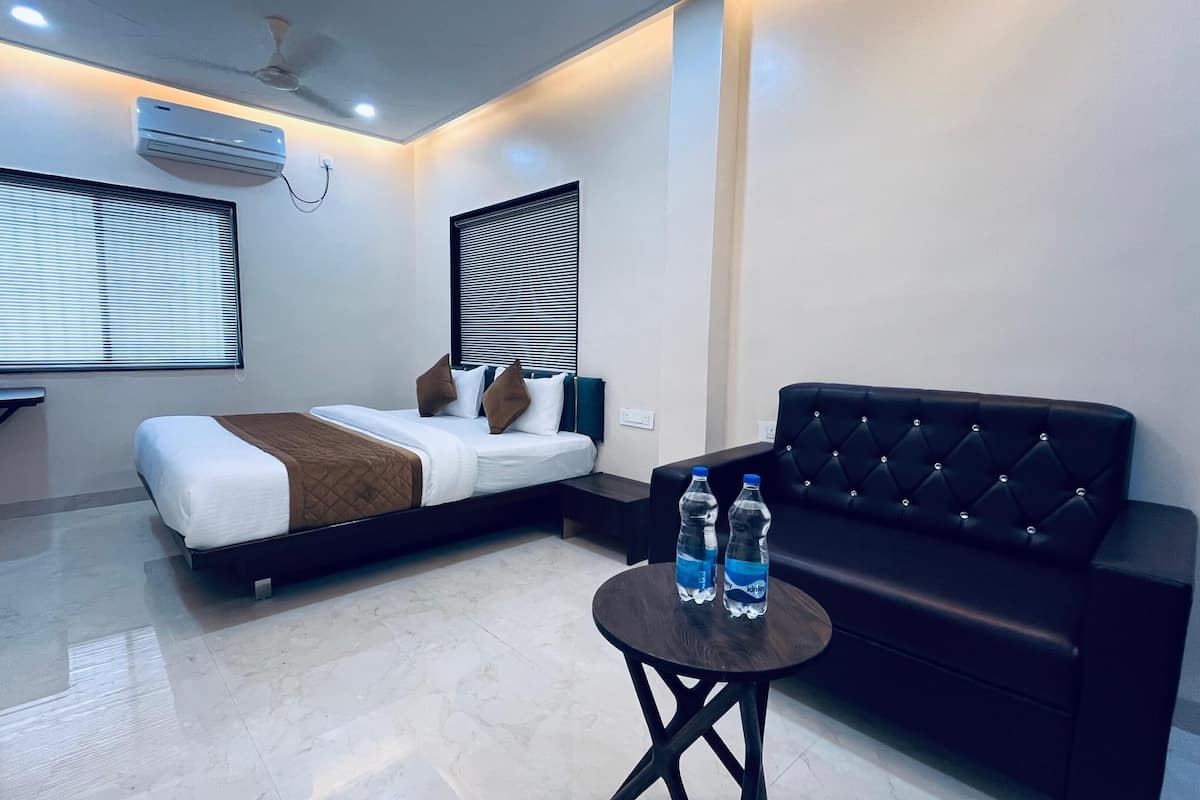 A modern hotel room with a bed, a black leather sofa, a small table with water bottles, and an air conditioning unit on the ceiling.