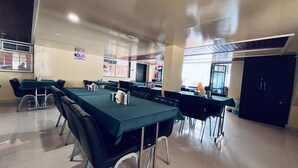 A spacious dining area with multiple tables and chairs, a visible kitchen area, and a large window providing natural light.
