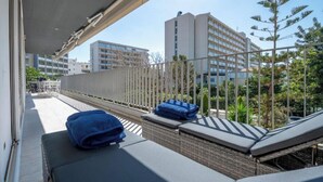 A balcony with sun loungers, a white fence, and a view of buildings and trees. - Athens Riviera Aelia Luxury Suite By the Sea - Location: Vouliagmeni, Kavouri  (Vouliagmeni)
