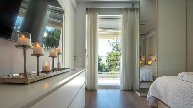 A hotel room with a bed, bedside table with candles, a large window with curtains, and a view of the outdoors. - Athens Riviera Aelia Luxury Suite By the Sea - Location: Vouliagmeni, Kavouri  (Vouliagmeni)