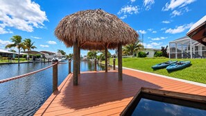 Una terraza de madera con techo de paja, un cuerpo de agua y kayaks sobre el césped.