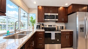 Una cocina moderna con armarios de madera oscura, electrodomésticos de acero inoxidable, encimera de granito y un amplio ventanal con vistas a una piscina y palmeras.