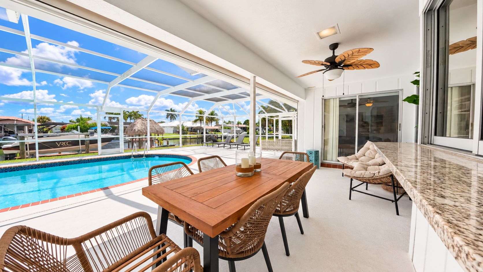 A poolside area with a wooden table and wicker chairs, a ceiling fan, and a view of the pool and resort.