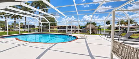 Un área de piscina con una piscina azul cristalina, una zona de estar techada y vistas a palmeras y edificios al fondo.