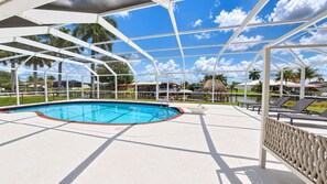 A pool area with a clear blue pool, a covered seating area, and a view of palm trees and buildings in the background. - VRCC Villa Sea Breeze - tropical retreat (Cape Coral)
