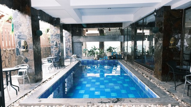 A swimming pool with blue tiles, surrounded by stone walls and white pebbles. There are chairs and tables around the pool, and plants are visible in the background. - Collection O Luna Hotel Pattaya (Pattaya)
