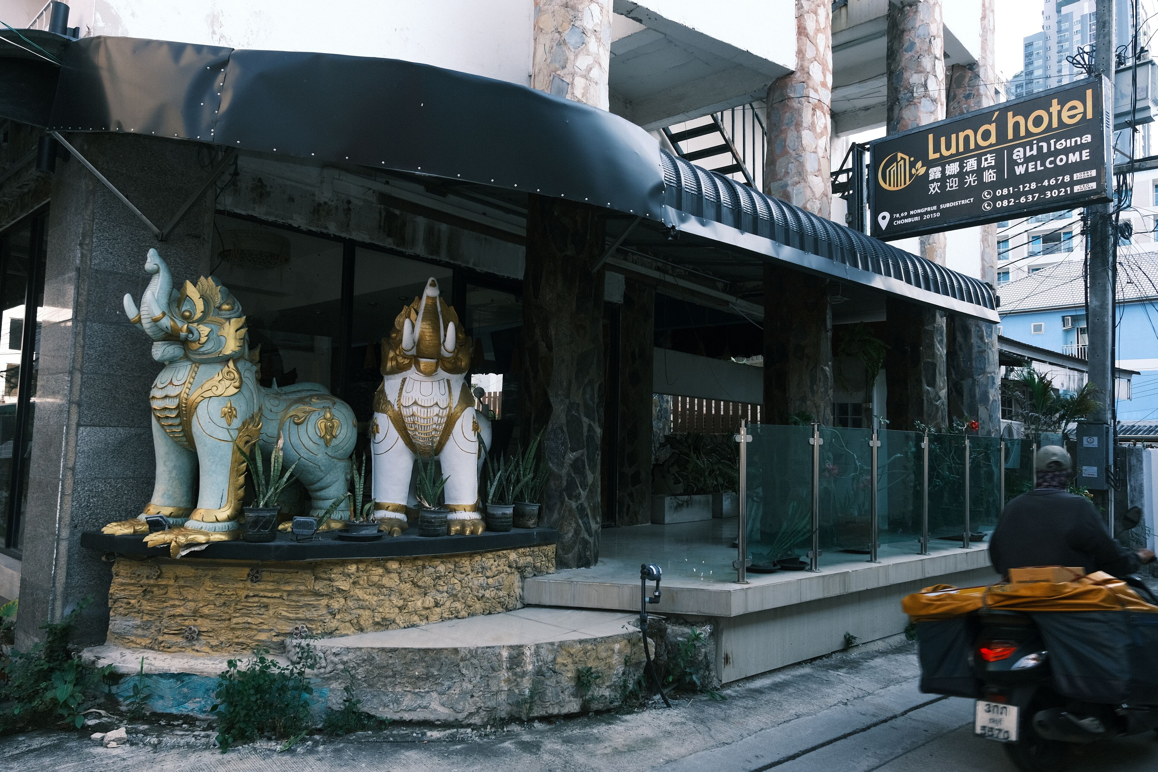 A hotel entrance with a signboard, a statue, and a motorcycle passing by.