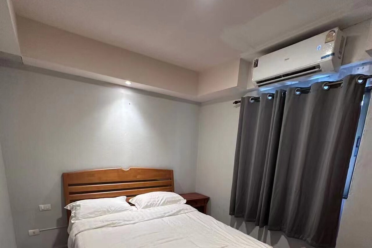 A bedroom with a wooden bed, white bedding, a marble-patterned floor, a nightstand, an air conditioning unit, and a window with grey curtains.