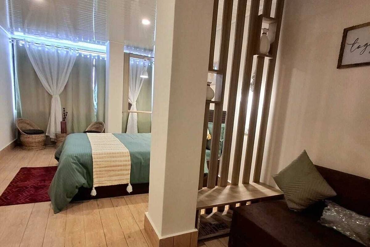A bedroom with a bed, a sofa, a window with curtains, a wooden floor, and a wall with a framed picture.