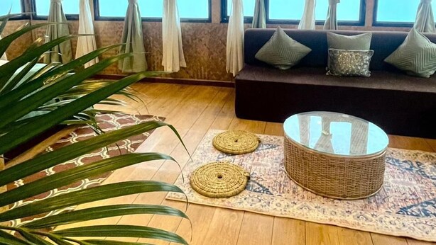A room with a wooden floor, a sofa with cushions, a round wicker table, and a large window with curtains. - Monratino Himadrie (Darjeeling)