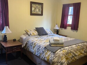 A neatly made bed with a patterned bedspread, a bedside table with a lamp, a framed picture on the wall, and a window with curtains. - Cozy Catskill Mountain Cabin (Margaretville)