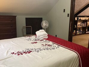 A bedroom with a bed, a fan, a red and white bedspread, a wooden dresser, and a door leading to another room. - Cozy Catskill Mountain Cabin (Margaretville)