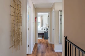 A hallway with a wooden floor, a wall-mounted decorative piece, and a bed in the background.