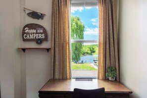 Ett rum med ett fönster som vetter mot en sjö, en skylt med texten ”GLADA CAMPARE” och en liten krukväxt.