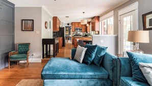 A modern living room with a blue sofa, wooden flooring, and a kitchen in the background. - Tranquil Family Retreat in NW Austin, Hot Tub, Office, & Backyard Escape (Austin)