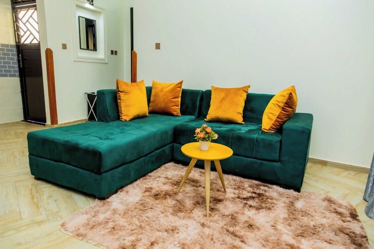 Un sofá seccional verde con cojines amarillos, una mesita redonda con un florero y una alfombra mullida sobre piso de madera.