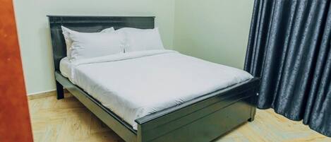 A single bed with a wooden frame and white bedding in a room with a herringbone-patterned floor and a black curtain.