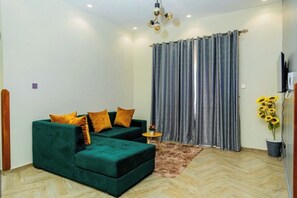 A living room with a green sofa, two orange cushions, a small round table, and a potted plant with yellow flowers. - Cozy Residences Kampala 003 (Kampala)