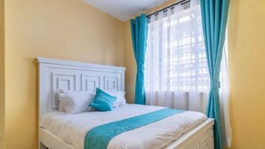 Una cama bien hecha con ropa blanca y una manta verde azulada, un cabecero blanco y una ventana con cortinas azules.