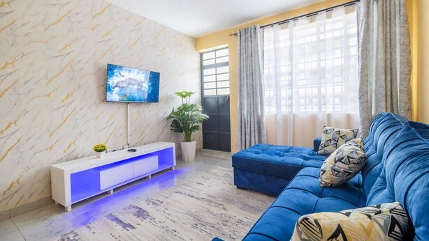 Una sala de estar con un sofá azul, un mueble blanco para entretenimiento con iluminación azul, un televisor grande en la pared y una planta.