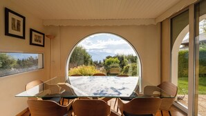 Un coin repas avec une table en verre et des chaises en bois, une grande fenêtre en arc de cercle offrant une vue sur la verdure et les montagnes, et des tableaux encadrés accrochés au mur.
