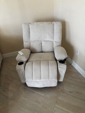 A beige reclining chair with a remote control and headphones on it, positioned against a wall with a tiled floor.