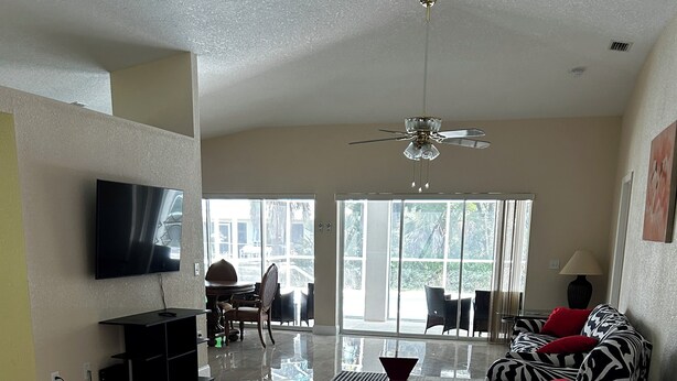A spacious living room with a flat-screen TV, a ceiling fan, and a large window offering a view of the outdoors.