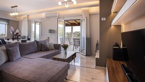 A modern living room with a sectional sofa, a coffee table, and a TV. There is a balcony with a view of the city.