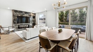 A modern living room with a marble wall, a flat-screen TV, a dining area with a wooden table and chairs, and a large window offering a view of the outdoors. - Gated,new 5bed home in Vegas Summerlin (Red Rock Casino area),w/ Hot Tub,BBQ,etc (Las Vegas)
