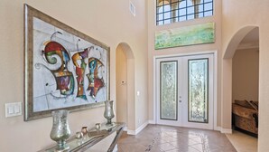 A spacious foyer with a modern art piece, a glass console table, and a tiled floor leading to a double door with glass panels.