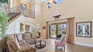 A spacious living room with a leather sectional sofa, a patterned armchair, and a glass coffee table. A staircase with a railing leads to an upper level.