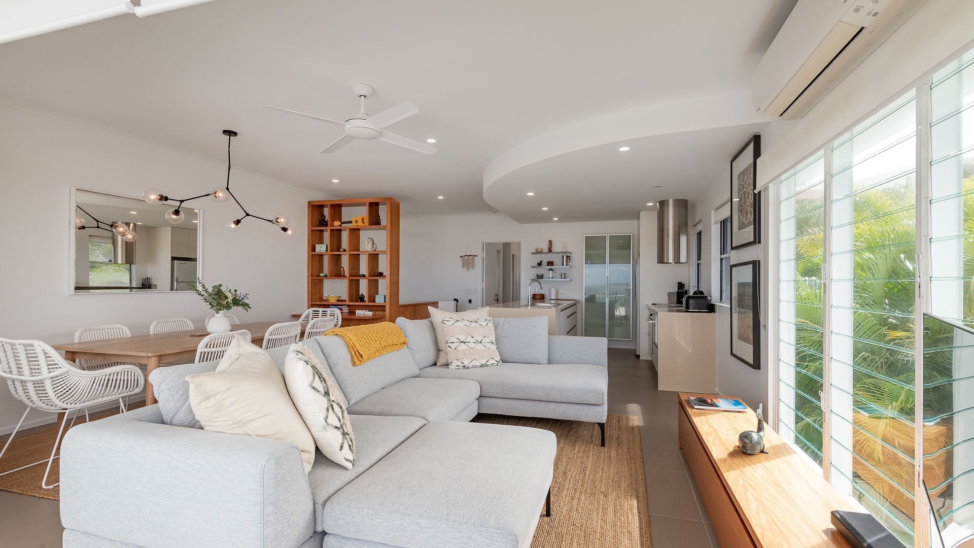 A modern living room with a grey sectional sofa, a dining area with white wicker chairs, a wooden bookshelf, and a kitchen with a glass door leading to a balcony.
