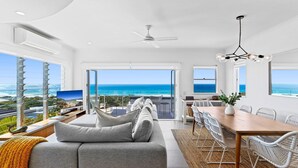 A modern living room with a large sofa, dining table, and chairs, overlooking a scenic ocean view.