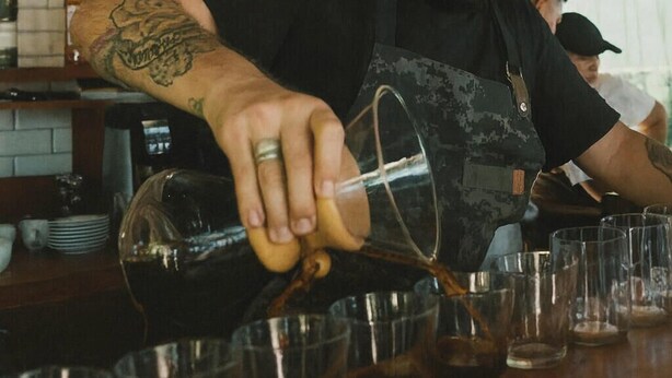 Un barista sirve café en vasos, con un letrero que anuncia recorridos de café y cacao.