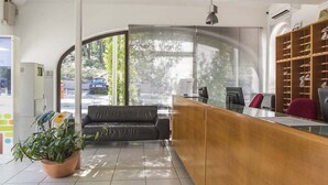 Un espace d’accueil avec un comptoir en bois, un canapé noir, une plante en pot et une grande fenêtre équipée de stores.