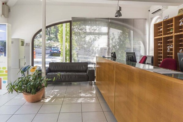 A reception area with a wooden counter, a black sofa, a potted plant, and a large window with blinds.