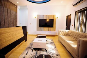 A modern living room with a flat-screen TV mounted on a wooden panel wall, a beige sofa, a wooden bench, and a patterned rug. - Vamble Apartments and Suites (Abuja)