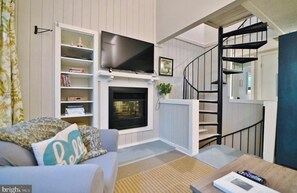 A living room with a fireplace, a TV mounted above it, a bookshelf, a staircase, and a cozy sofa. - Spectacular Treehouse - Lake, Firepit + FREE Wine (Basye)